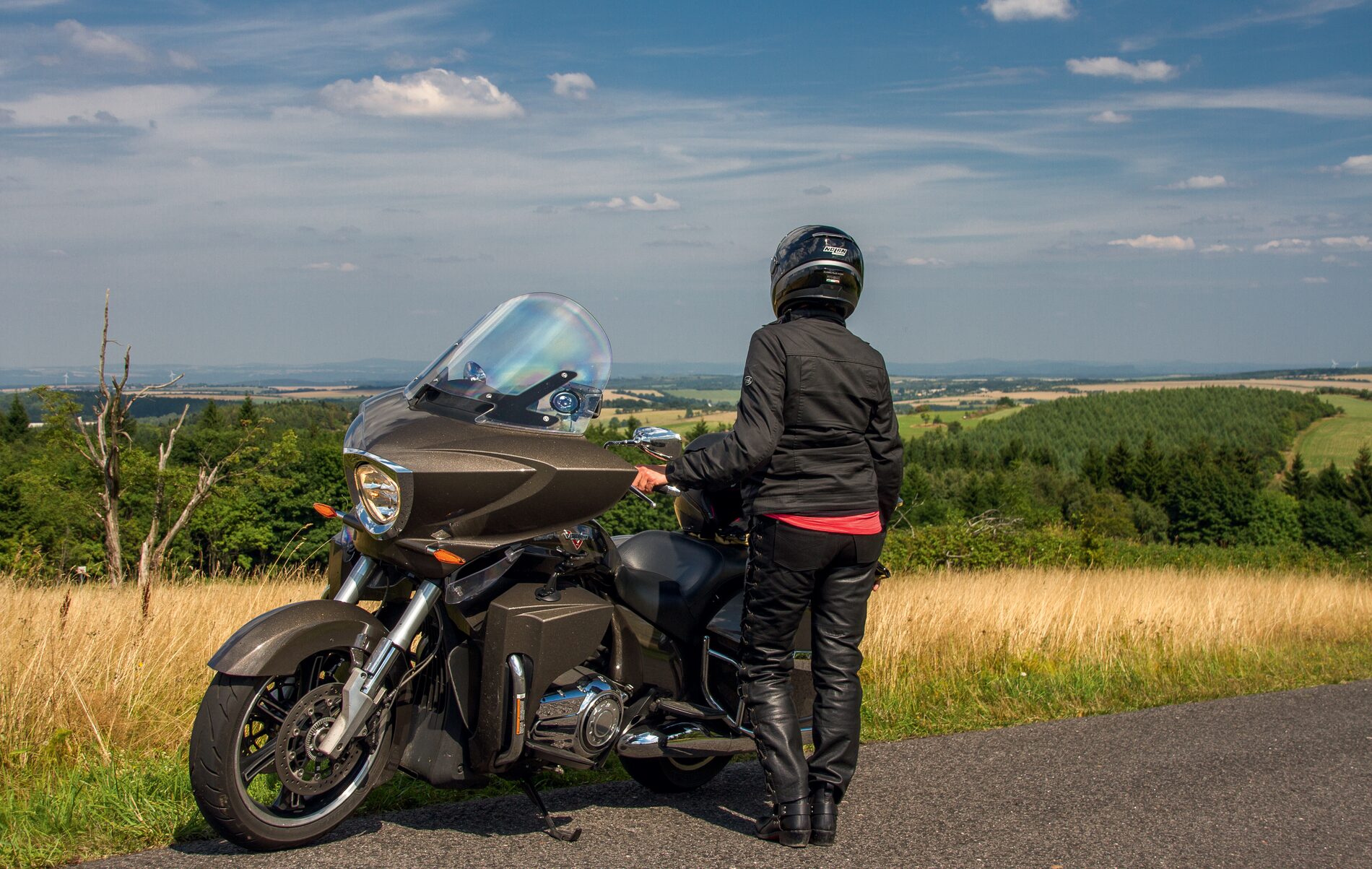 Motorradfahrerin neben ihrer Maschine an einem Straßenrand mit weitem Blick über Wiesen und Wälder